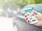 Rideshare taxi app on smartphone screen. Online ride sharing and carpool mobile application. Modern people and commuter transportation service. Man holding phone with a car in background.